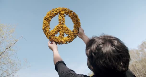 Un hombre sostiene en la mano un gran símbolo de paz hecho de flores amarillas en el cielo azul — Vídeos de Stock