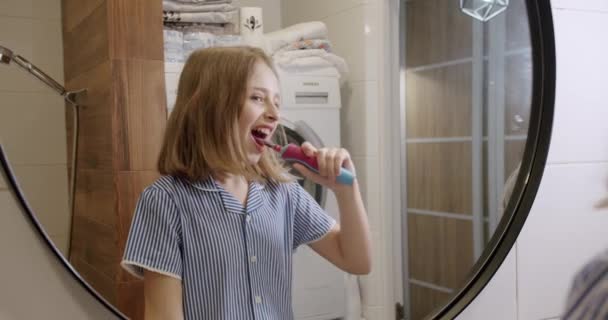 A teenage girl brushes her teeth with an electric toothbrush in the bathroom — Stockvideo