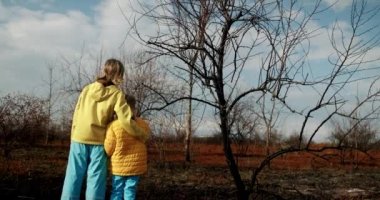 The younger sisters dressed in the colors of the Ukrainian flag embrace and encourage each other on a background of earth burned during the war. Stop the war in Ukraine. homeless child. No War.