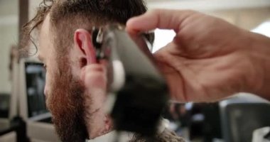 Yakışıklı adam elektrikli budayıcı ile yeni bir saç stili alıyor. Makyaj işiyle kapat. Modern berber dükkanında sakal ve bıyık bakımında genç hippi beyaz adam. Erkek saç stili. 