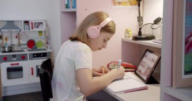 The teenager is at home drawing at the table, the childrens room in pink colors. Take homework lessons. Teenager with headphones writes on notebook and uses tablet gadget for internet.