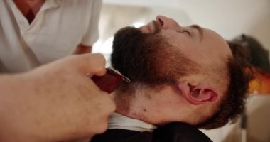 Yakışıklı adam elektrikli budayıcı ile yeni bir saç stili alıyor. Makyaj işiyle kapat. Modern berber dükkanında sakal ve bıyık bakımında genç hippi beyaz adam. Erkek saç stili. 