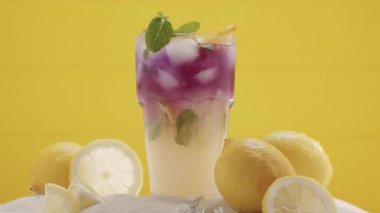Buz ve limon parçaları ferahlatıcı yaz limonata kokteylinin içine düşer, yaz ferahlatıcı kokteyl, sarı arka planda deniz kumu ve tropikal yapraklarla çekilmiş bir stüdyo çekimi.