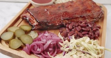 İçinde kirazlı chipotle soslu bebek kaburgaları olan büyük bir tabak. Sigara, sos, barbekü fasulyesi, lahana salatası, soğan turşusu, salatalık turşusu, izole edilmiş beyaz arka planda dönerek yavaş yavaş pişiyor.