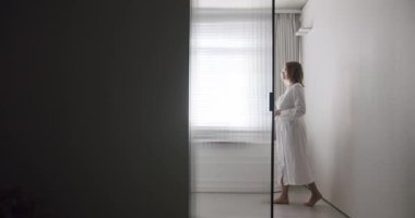Woman in robes open Transparent sliding door inside the modern bathroom, minimalist bathtub with wood accessories in white and white colors. Modern Minimalist White Bathroom. white minimalist interior