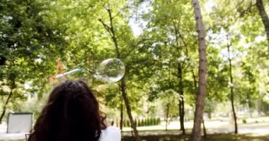 Fashionable young caucasian woman blowing soap bubbles in the nature. Young One Woman Smiling Carefree Having Fun Blowing. Joy Girl Enjoying Holidays. Young woman making large soap bubbles and smiling