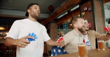 Happy mixed race supporters cheering and exulting after winning. Friends at the bar in a pub having fun. Men with the flag of the national country of USA drinking beer and watching sport game.