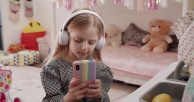 7 yaşındaki kız çocuk odasında beyaz kulaklıklarla oynuyor ve renkli akıllı telefon kullanıyor. Sosyal ağda havalı video veya fotoğraf içeriğinin keyfini çıkarmak, çevrimiçi oyunlar oynamak.