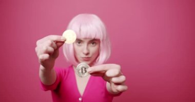 Pembe saçlı güzel bir kadın, elinde bir bitcoin tutuyor, onu yakından gösteriyor, kripto para sembolü. Gelecekteki para birimi. Pembe arka planda kapalı stüdyo çekimleri. Yeni Sanal Para.