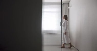 Woman in robes open Transparent sliding door inside the modern bathroom, minimalist bathtub with wood accessories in white and white colors. Modern Minimalist White Bathroom. white minimalist interior