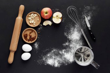 Siyah masada turta yapmak için malzemeler ve mutfak aletleri var: un, yumurta, elma, merdane, çırpı, süzgeç..