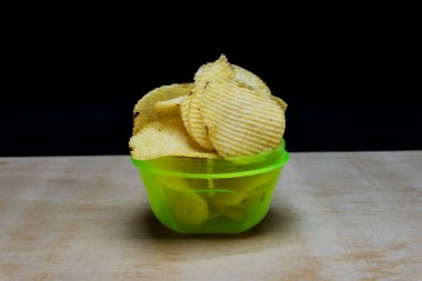 Masada yeşil kase içinde tuzlu patates cipsi gofretleri, siyah arka planda izole edilmiş cips parçaları.