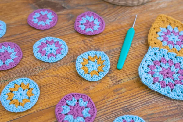 Side view of multicolored round crochet patterns in selected focus, blurred crochet hook and square motif on wooden scratched background. Crocheting as a hobby concept.