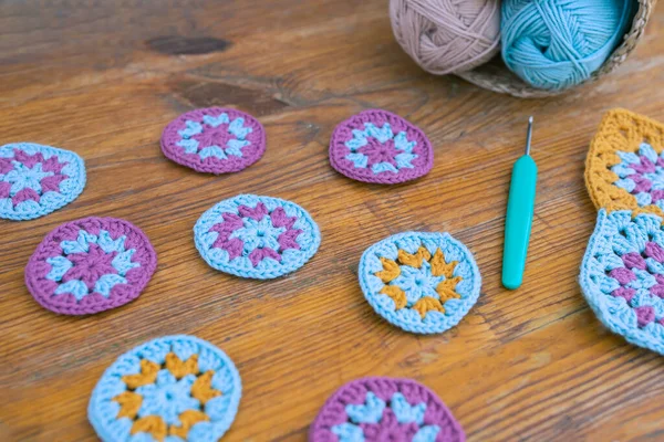 Side view of small round crochet patterns with selected focus on wooden surface, blurred crochet hook and skeins of yarn lying in the background. Crocheting multicolored patterns concept.