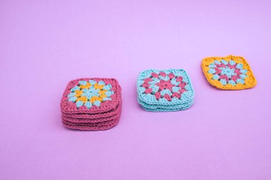 Three piles of square crochet motifs, sorted diagonally from tallest to smallest on light violet smooth background. Crocheting granny square motifs concept.