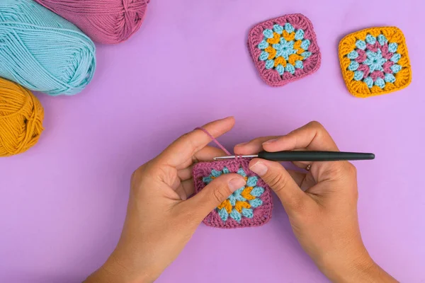 Top view of wonan's hands crocheting multicolored granny square with skeins of cotton yarn and finished squares on the background. Craft work concept.