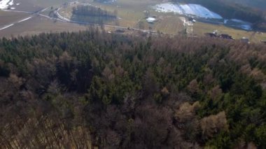 Köyün yukarısında yoğun ormanlar, evler, tarlalar, saman balyaları ve park etmiş turistlerle dolu bir tepe.