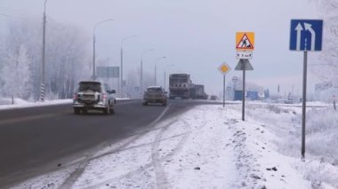 Kış. Yoldaki araba trafiği. Mavi gökyüzü. Kirli kar. Farları açık arabalar. Yol boyunca aydınlatma direkleri var. Yol siyahtır. Gri kaldırım. Yol işaretleri. reklam panoları