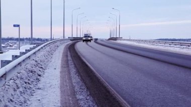 Kış. Yoldaki araba trafiği. Mavi gökyüzü. Kirli kar. Farları açık arabalar. Yol boyunca aydınlatma direkleri var. Yol siyahtır. gri kaldırım.