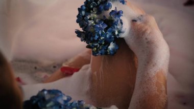 Köpük banyosunda rahatlayan rahat bir kadının yakın çekimi. Bir kadın mavi bir ortanca çiçeğiyle tenine dokunuyor..
