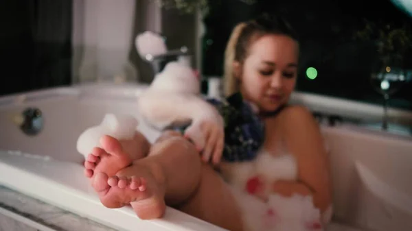 Close-up de uma mulher relaxada relaxante em um banho de espuma. Uma mulher toca sua pele com uma flor de hortênsia azul. — Fotografia de Stock