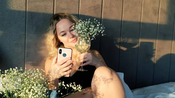 Mutlu ve şehvetli bir kadın evde yatarken cep telefonuyla çiçeklerle selfie çekiyor.