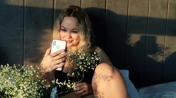 Mutlu ve şehvetli bir kadın evde yatarken cep telefonuyla çiçeklerle selfie çekiyor.