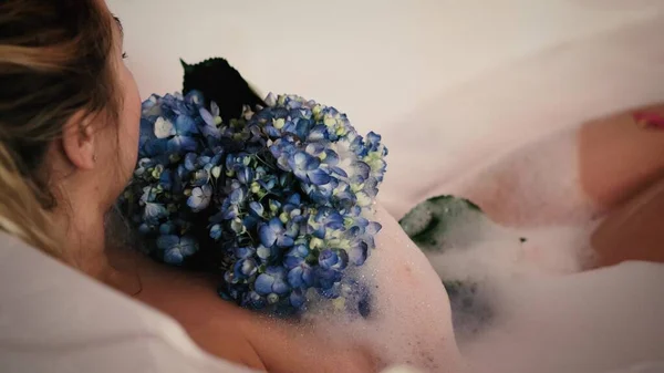 Close-up de uma mulher relaxada relaxante em um banho de espuma. Uma mulher toca sua pele com uma flor de hortênsia azul. — Fotografia de Stock