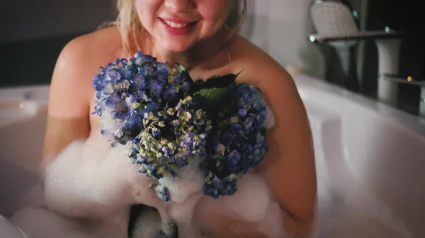 Primer plano de una mujer relajada relajándose en un baño de burbujas. Una mujer toca su piel con una flor de hortensia azul. — Foto de Stock