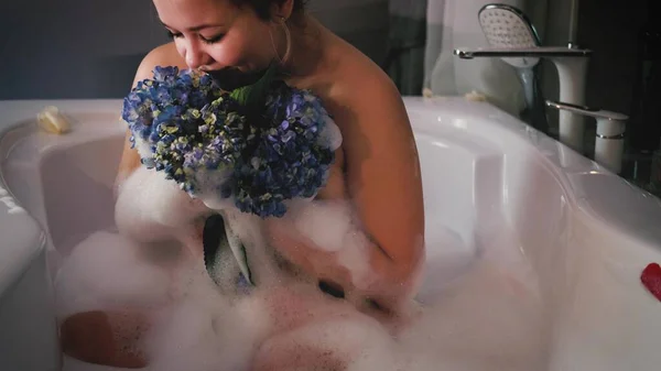 Nahaufnahme einer entspannten Frau, die sich in einem Schaumbad entspannt. Eine Frau berührt ihre Haut mit einer blauen Hortensienblüte. — Stockfoto
