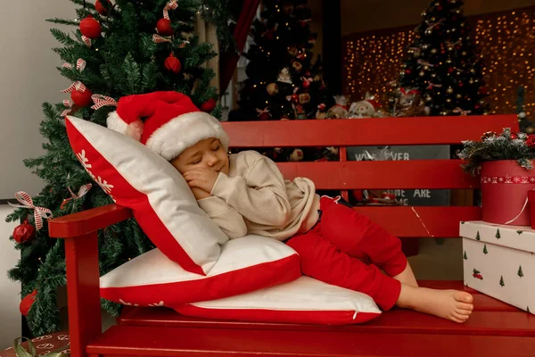 Noel Baba 'lı bir çocuk Noel ağacının yanında uyur ve gülümser.
