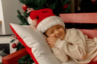 Noel Baba 'lı bir çocuk Noel ağacının yanında uyur ve gülümser.