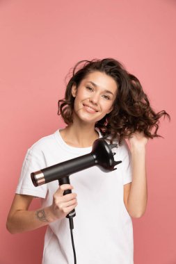 Saç kurutma makinesi ve pembe arka planda uzun kablosu olan kıvırcık kadın portresi