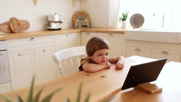 Un poco de chico caucásico come crujiente y mira tableta PC en la mesa de la cocina. — Vídeo de stock