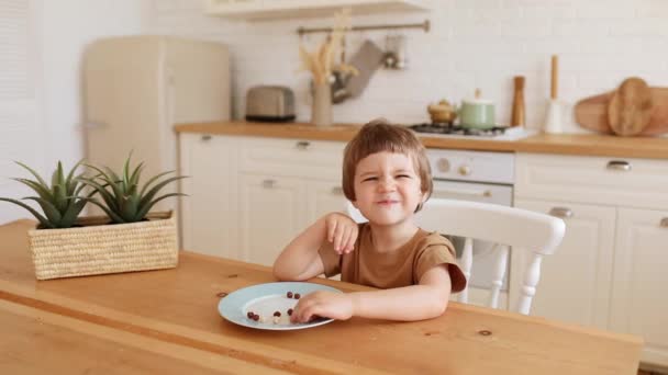 Un pequeño chico caucásico come un desayuno crujiente y lo disfruta en la mesa de la cocina. — Vídeo de stock