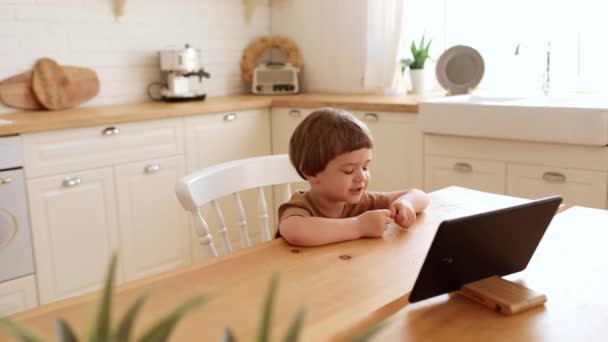 Egy kis kaukázusi fiú eszik krunchy és néz tabletta PC a konyhaasztalnál. — Stock videók