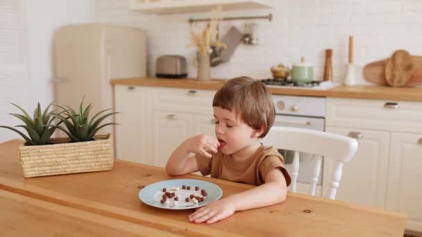 A little caucasian boy eats krunchy breakfast and enjoys it at kitchen table. — Stock Video