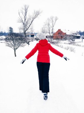 Kırmızı ceketli, beyaz şapkalı ve siyah eşofmanlı bir kadının kışın şehrin dışında karda yürümesi.