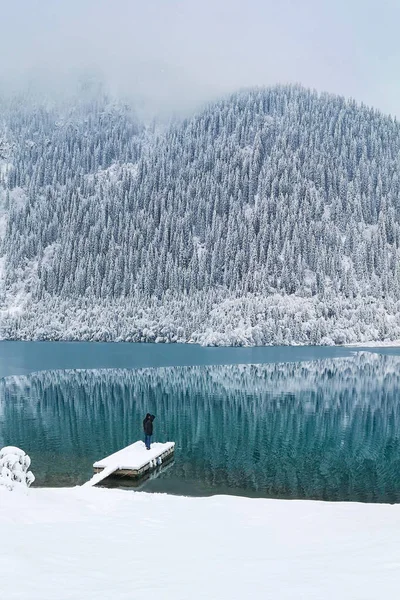 A man stands on the boat pier of a mountain lake in winter