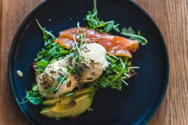 Kahvaltı için restorandaki siyah tabakta roka, somon ve avokado ile Benedict usulü yumurta..