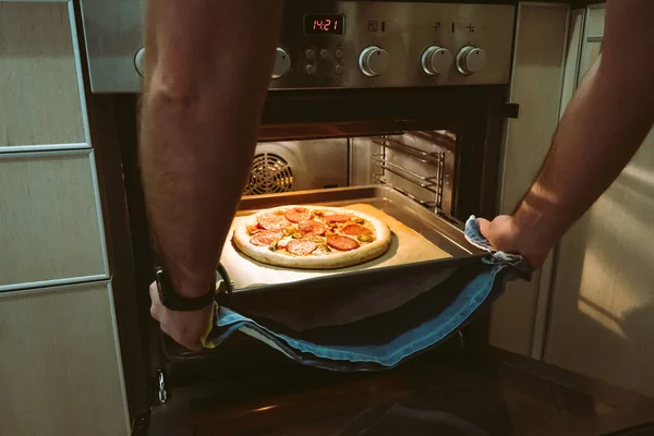 A man bakes pizza at home using an oven. Mens hands pull a pizza baking sheet out of the oven