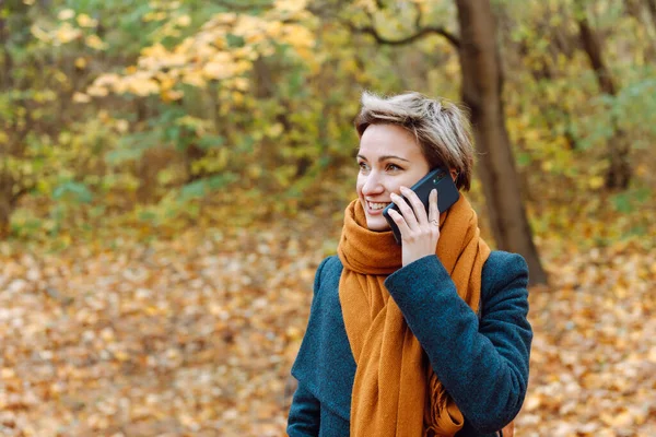 Short-haired blonde smiles as she talks on the phone in the fall on the street.