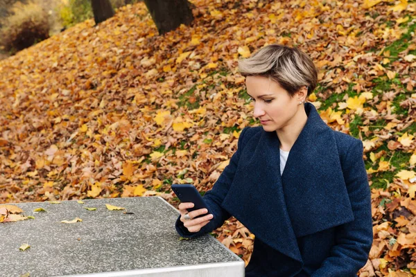 Short-haired blonde uses a smartphone sitting at a table in the park in the fall.