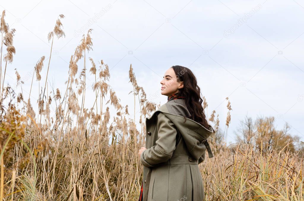 Young dreamy woman wearing grey old fashioned coat stands in the dry grass reeds field looking away.