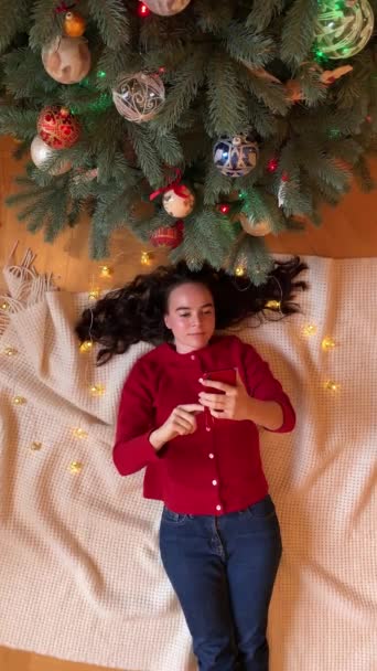 Vista dall'alto di una bella giovane donna sdraiata vicino all'albero di Natale, che sorride, tiene in mano e fa scorrere il suo telefono. — Video Stock