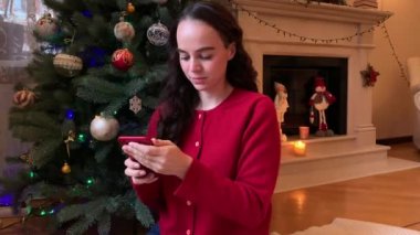 Genç bir kadın akıllı telefonu kaydırıyor, Noel hediyesi alıyor, Noel ağacı ve şöminenin yanında oturuyor..