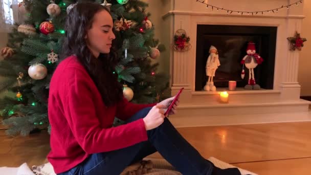Молода жінка прокручує смартфон, купує різдвяні свята онлайн, сидячи біля ялинки та каміна.. — стокове відео