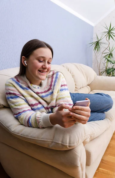 Joyeux rire adolescent fille textos, vidéo appelant sur son smartphone tout en se relaxant à la maison sur le canapé. — Photo