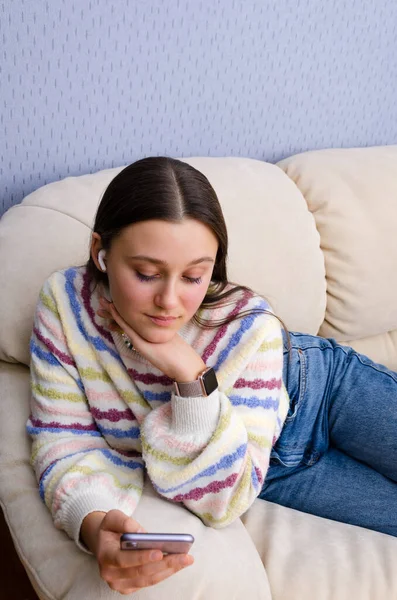 Adolescente rêveuse se relaxant sur un canapé, en utilisant smartphone regarder la vidéo dans les écouteurs. Écouter de la musique en ligne. — Photo