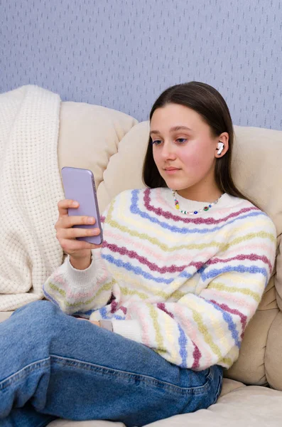 Портрет молодої дівчини-підлітка, яка використовує смартфон з навушниками в соціальних мережах, перегляд відео, розслабляється на дивані . — стокове фото